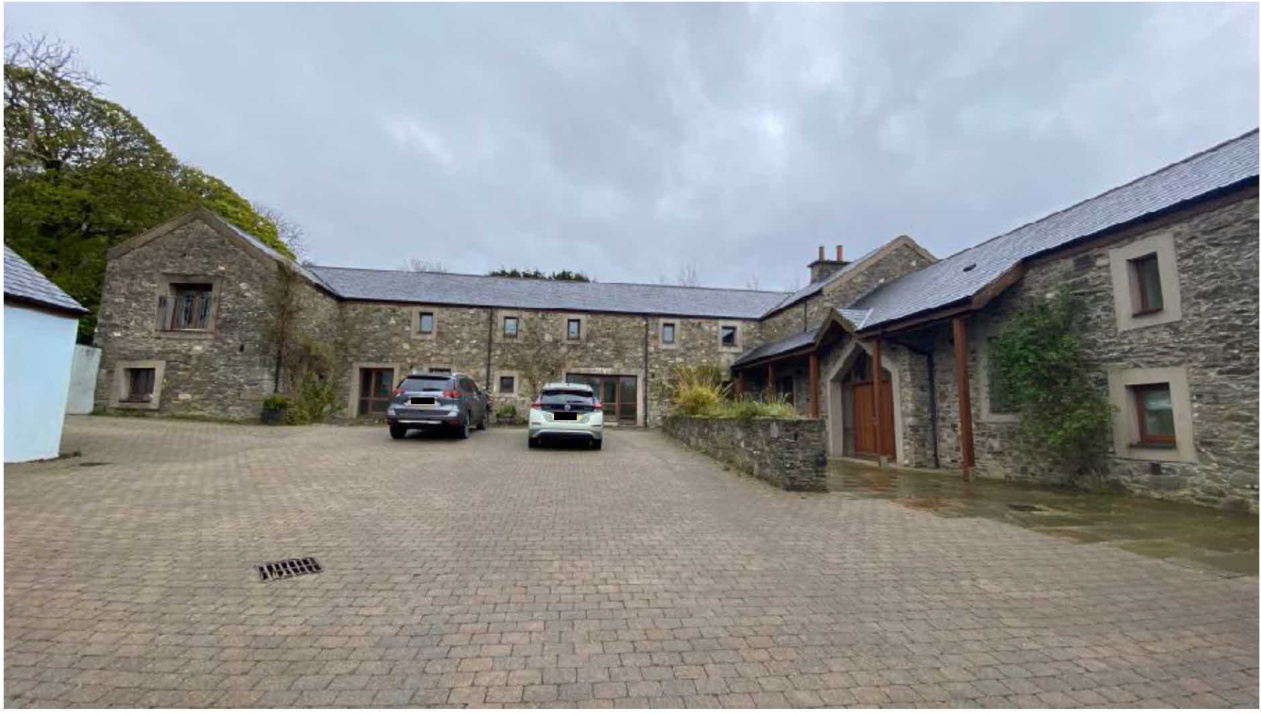 A photograph showing a large stone property complex, likely a converted barn, with a paved driveway and parked cars.