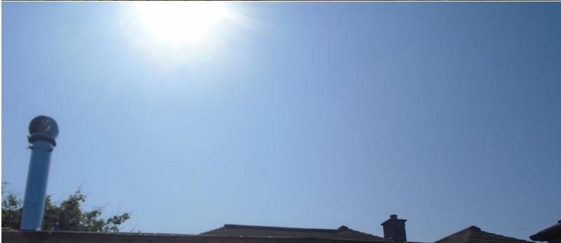 A low-angle photograph showing a bright blue sky with the sun, featuring the roofline of a residential property with a distinctive blue chimney pot and brick chimneys.