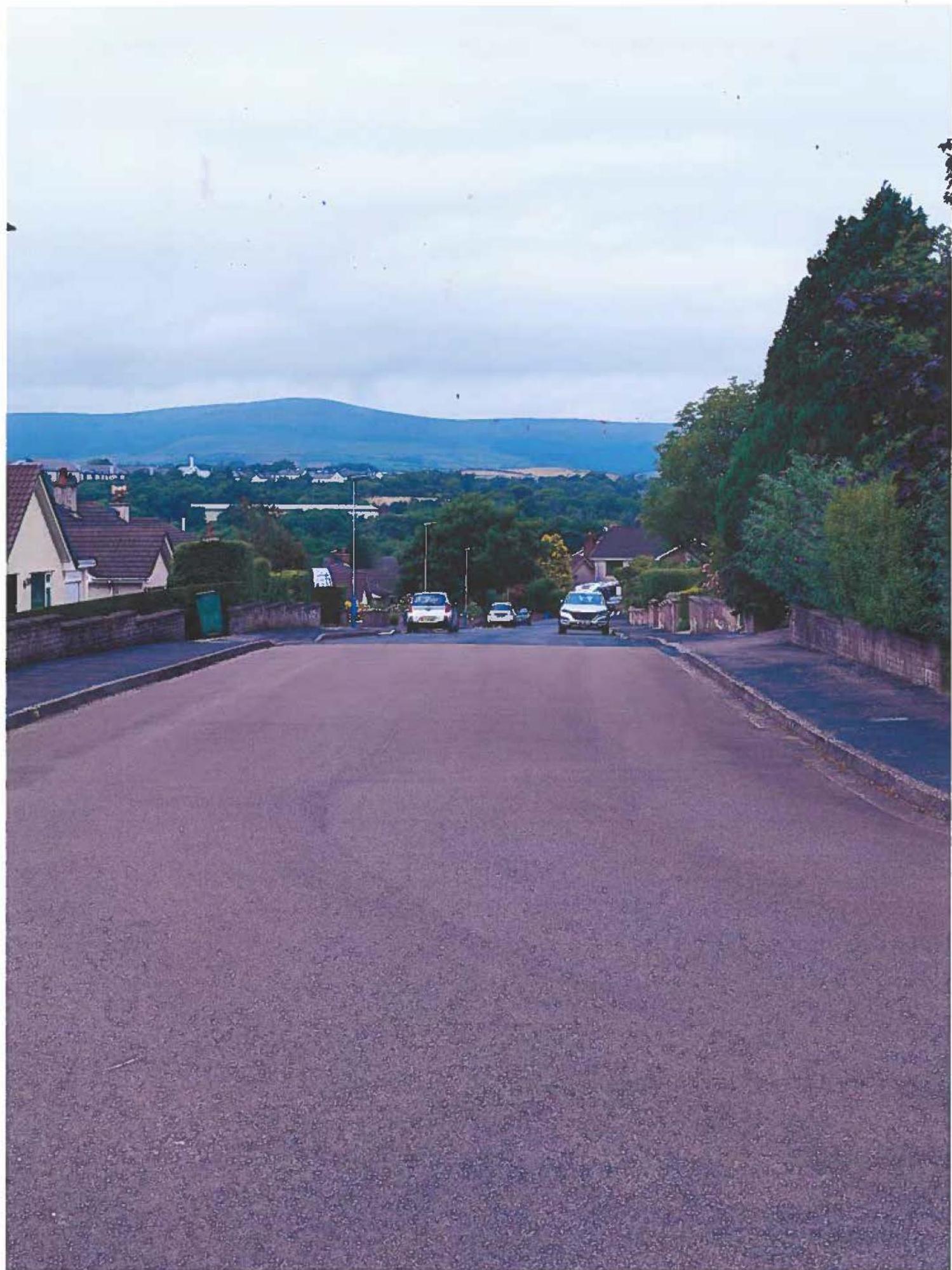 A photograph showing a paved residential road leading towards a valley with hills in the distance, flanked by houses and trees.