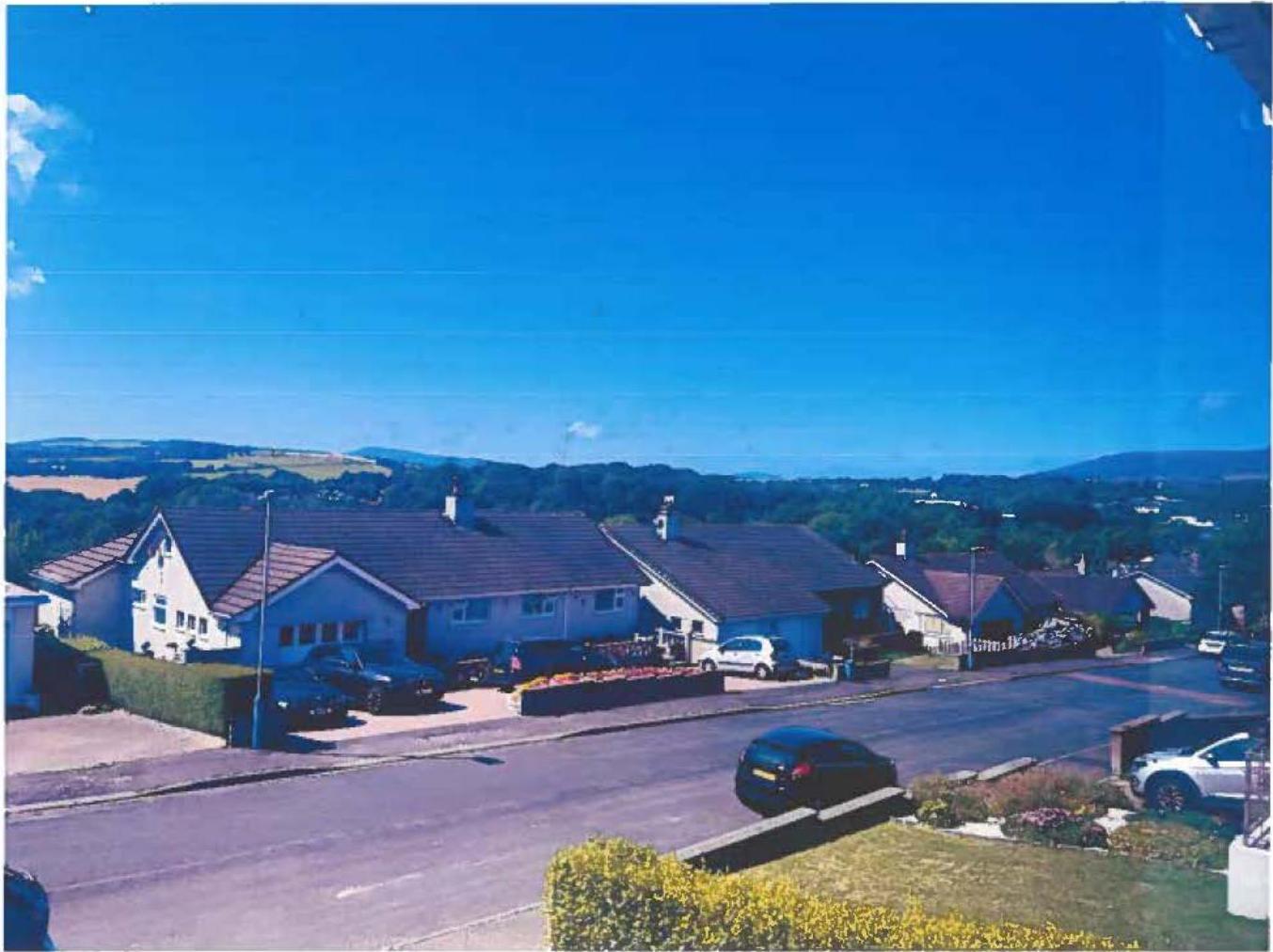 A photograph showing a residential street with detached bungalows and parked cars, set against a backdrop of green hills under a clear blue sky.