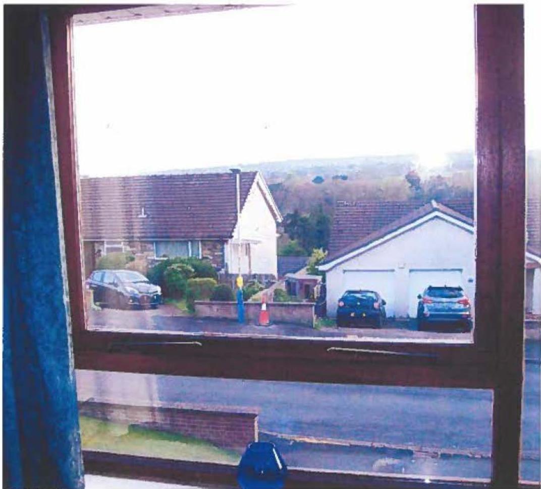 A photograph taken from inside a property looking out through a window frame at a residential street with houses and parked cars. The view captures neighboring properties including a white garage and a stone house.