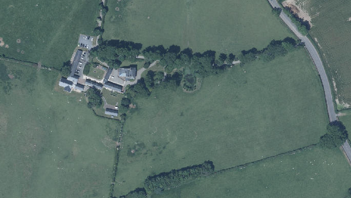 An aerial view of a rural property featuring a cluster of buildings surrounded by green fields, hedgerows, and a nearby road.