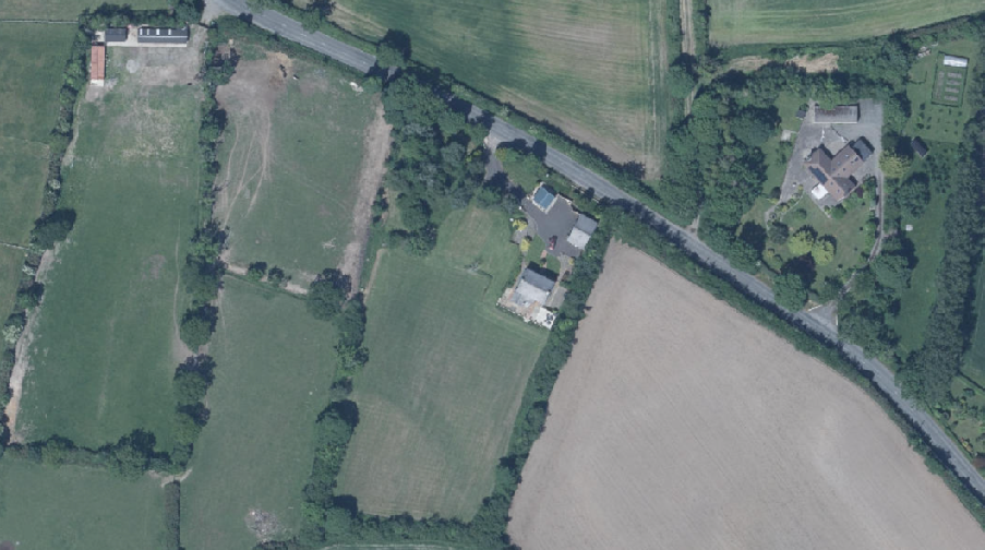 Aerial satellite view of a rural property site showing fields, hedgerows, and nearby residential buildings along a road.