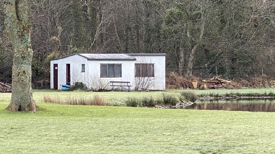 A photograph of a white single-story building, likely a workshop, situated in a grassy area near a small pond and wooded background.