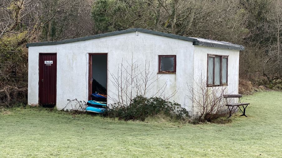 A photograph showing the exterior of a white single-story outbuilding or workshop situated in a grassy area with trees in the background.