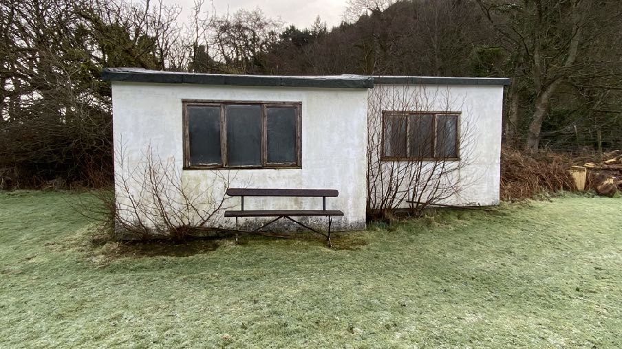 A photograph of a white, single-story outbuilding or workshop situated in a grassy, rural area with trees in the background.