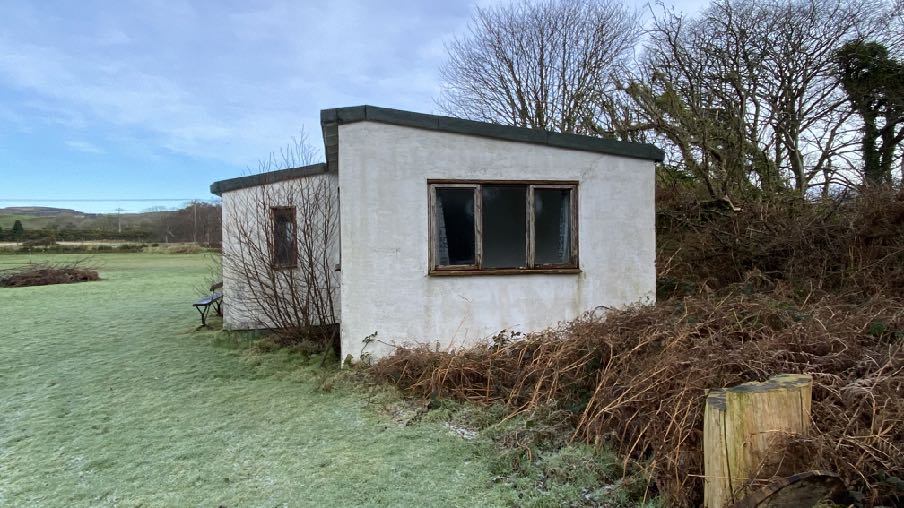 A photograph of a small, white, single-story outbuilding or workshop situated in a grassy rural area with overgrown vegetation nearby.