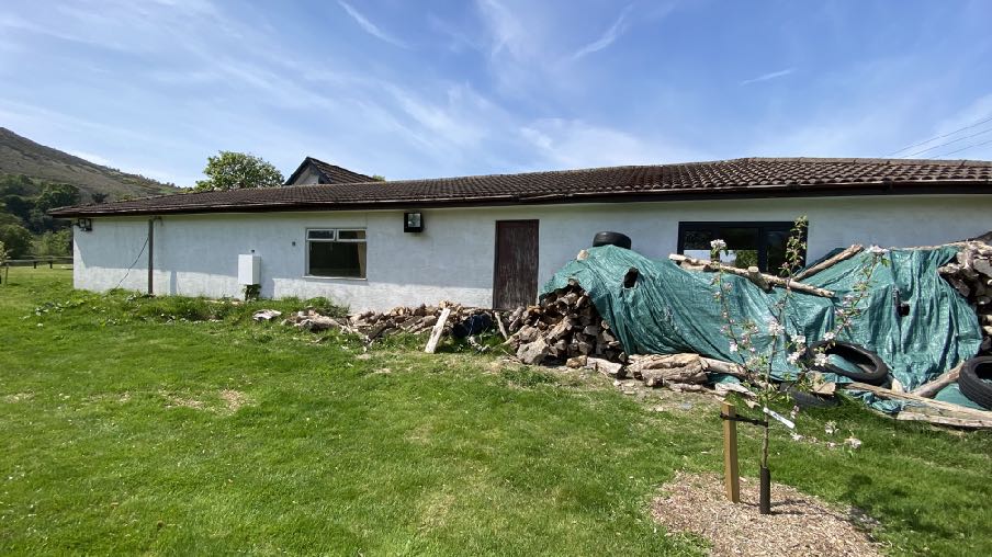 A photograph showing the exterior of a single-story white building, likely a workshop, with a grassy yard and a pile of wood covered by a tarp.