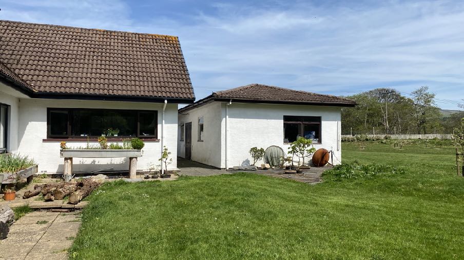 A photograph showing the exterior of a white bungalow and an attached single-story workshop or outbuilding with a tiled roof, set against a grassy garden.