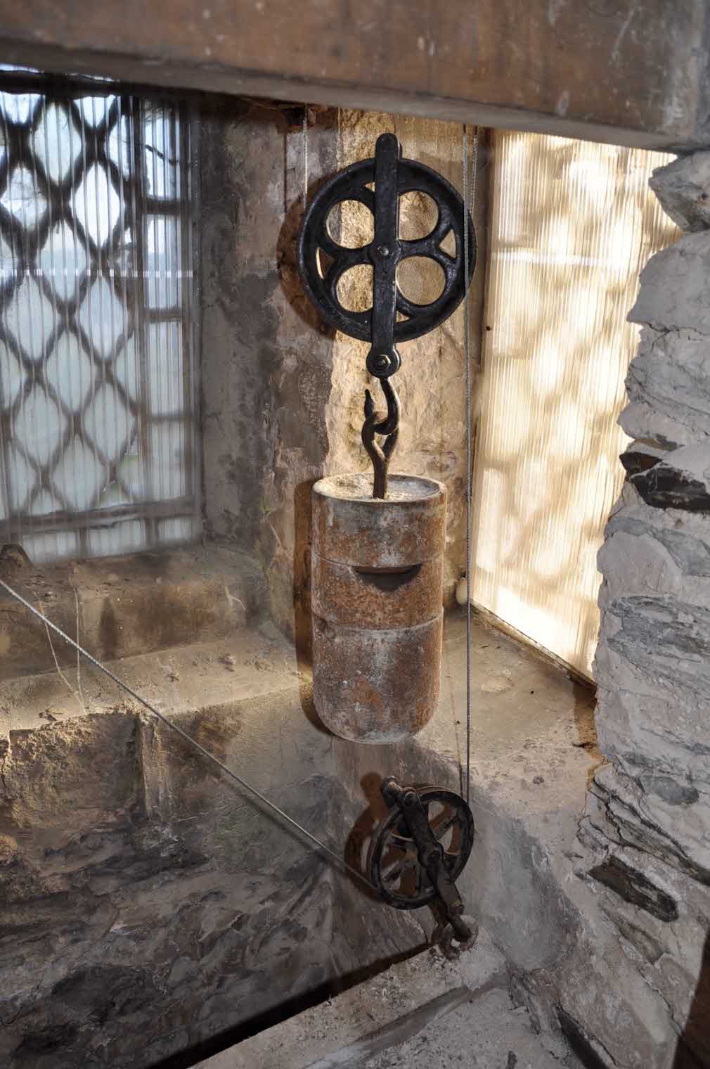 An interior photograph of a stone building showing a heavy metal weight suspended from a pulley system next to a diamond-paned window.