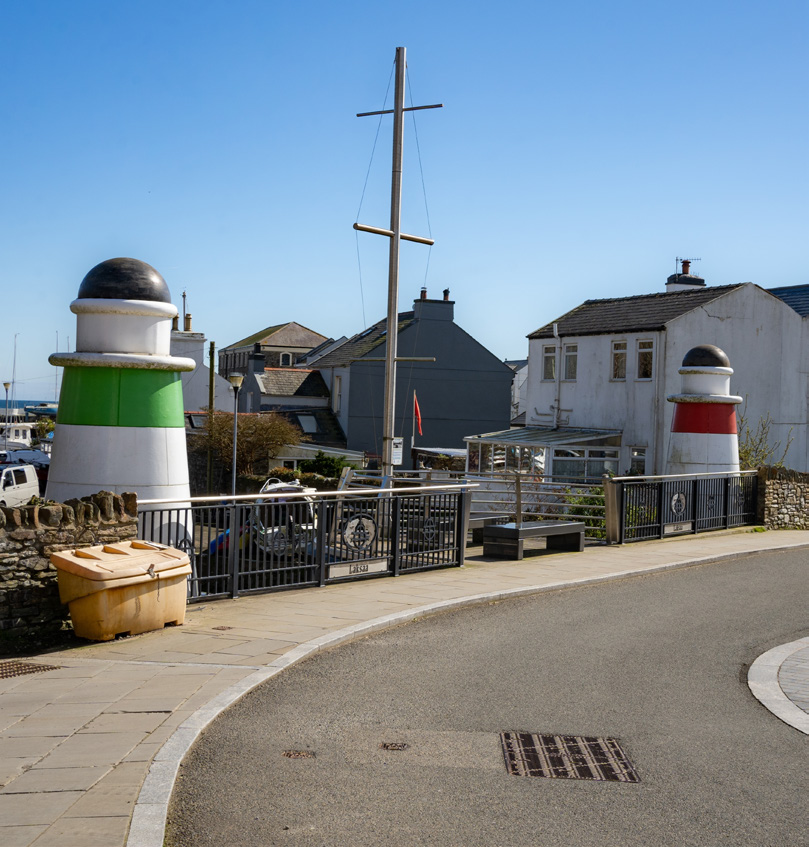 A photograph showing a paved road curving past decorative lighthouse pillars and a railing, with residential buildings and a mast in the background.