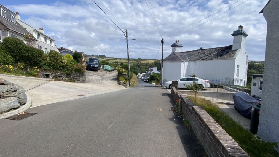 A street-level photograph showing a sloping residential road in a village setting with white houses, parked cars, and stone boundary walls.