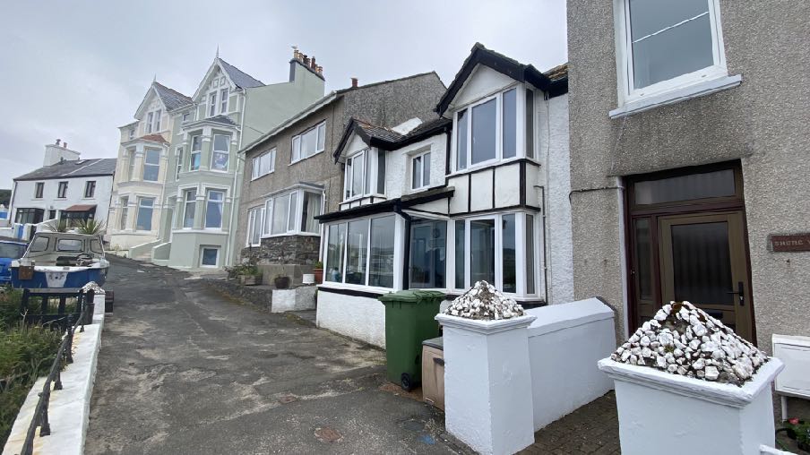A street-level photograph showing a row of residential properties in a coastal village, featuring a house with a black and white timber facade and a large conservatory extension.