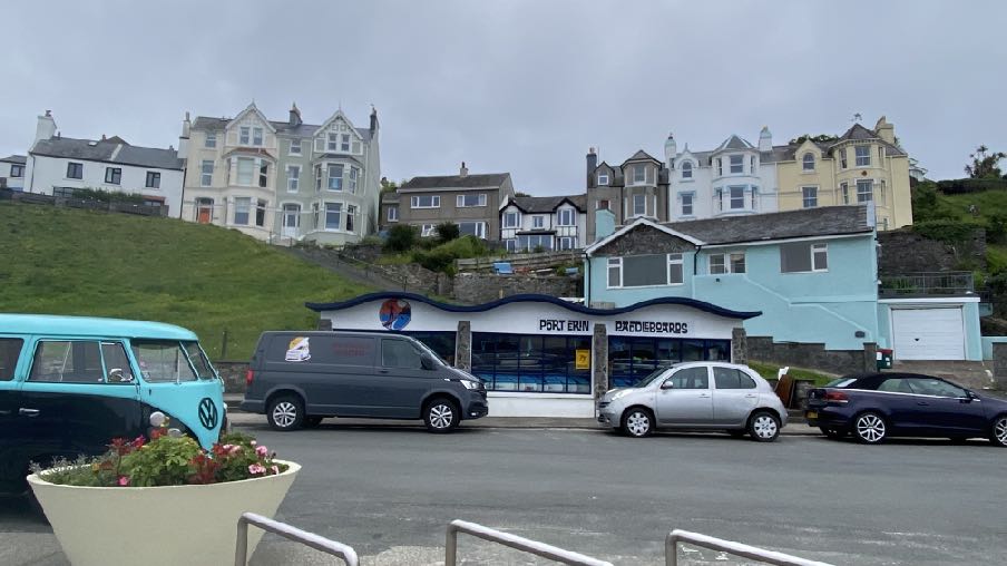 A street-level photograph showing a parking area with vehicles in front of a shop, with a grassy hill and large residential houses in the background.