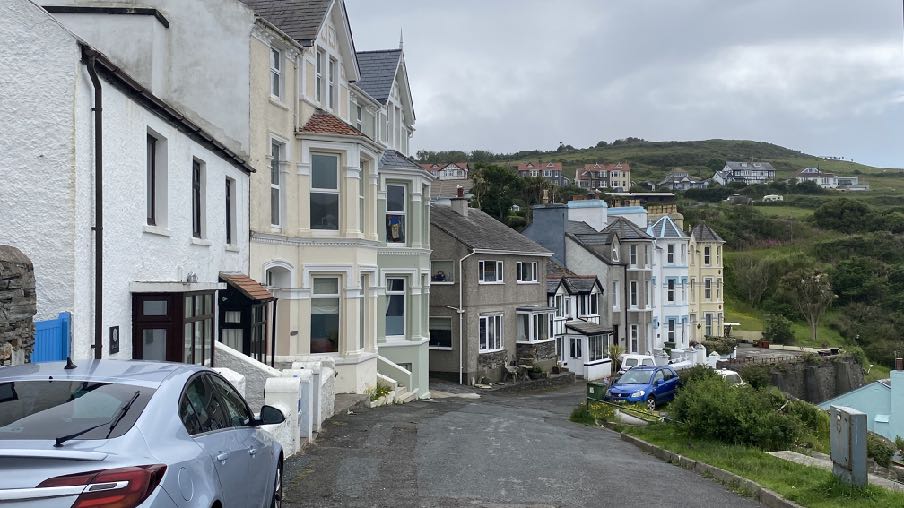 A street-level photograph showing a row of residential houses on a hillside in a coastal town, with a car parked in the foreground.