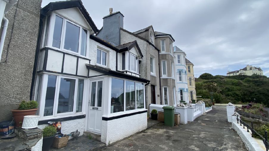 Street-level photograph of a white and black timber-framed terraced house with a large bay window and porch, situated in a row of similar buildings.