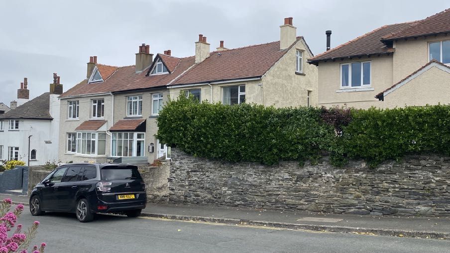 A street-level photograph showing a row of terraced houses with pitched roofs and chimneys, separated from the road by a stone wall and hedge.