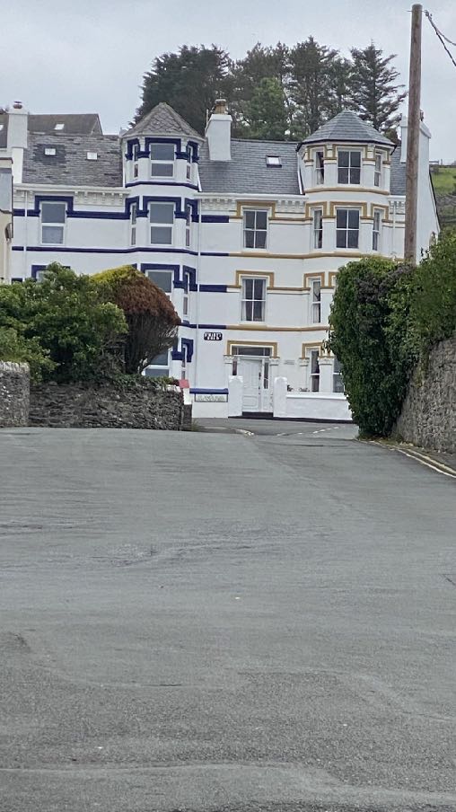 A photograph showing a large, white multi-story building with blue and gold trim situated on a hillside, viewed from a paved road.