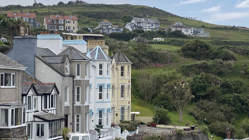 A photograph showing a row of terraced houses in the foreground with a grassy hillside and scattered residential properties in the background.