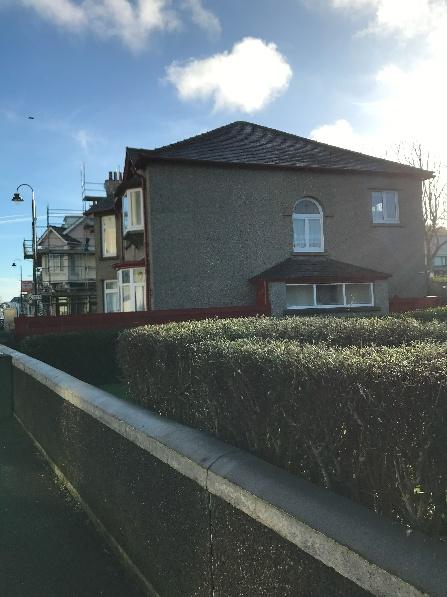 A street-level photograph of a two-story residential building with pebbledash rendering and a tiled roof, viewed from behind a hedge and low wall.