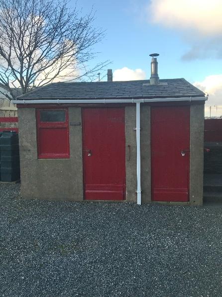 A photograph of a small, single-story concrete outbuilding or garage featuring a slate roof and two large red doors.