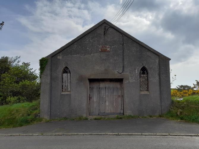 A photograph of a grey, single-storey building with a gabled roof and arched windows, identified as a former chapel. It is situated on a grassy verge next to a road.