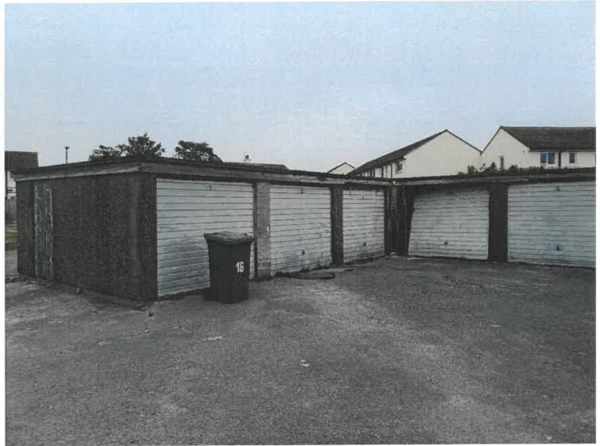 A black and white photograph showing a row of disused garage blocks with roller shutter doors and a wheelie bin in the foreground.