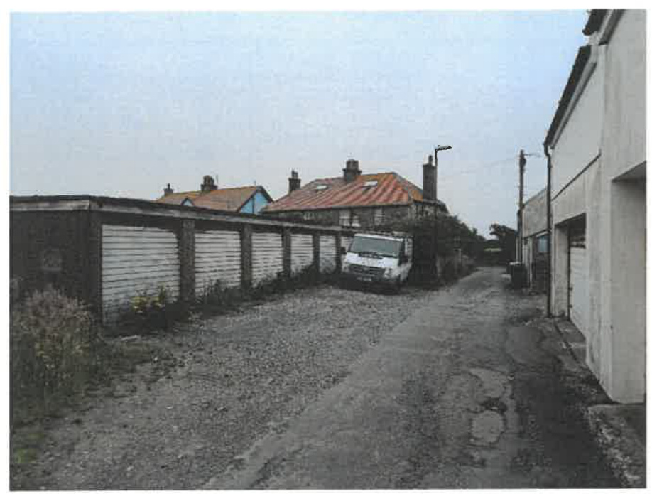 A photograph showing a row of single-bay garages with white roller doors along a gravel lane, with a white van parked in front and houses in the background.
