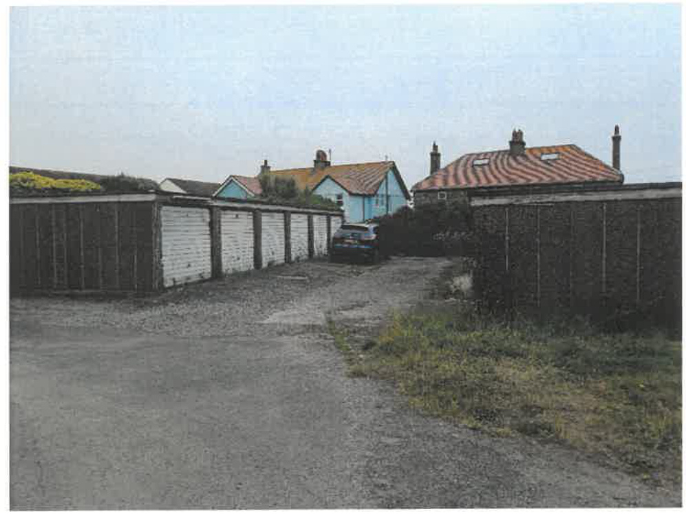 A photograph showing a row of garage blocks with white doors along a driveway, with residential houses visible in the background.