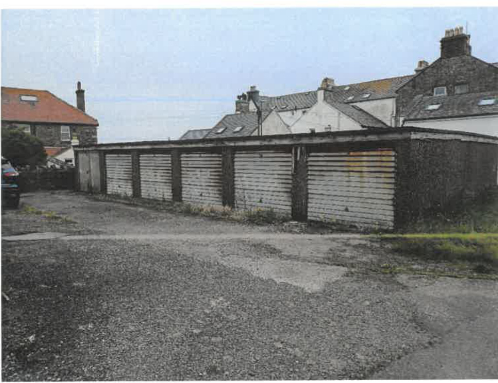 A photograph showing a row of disused garages with corrugated doors, situated in front of residential houses with slate roofs.