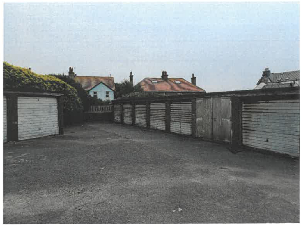 A photograph showing a row of disused garages with roller doors and a paved forecourt, with residential houses visible in the background.