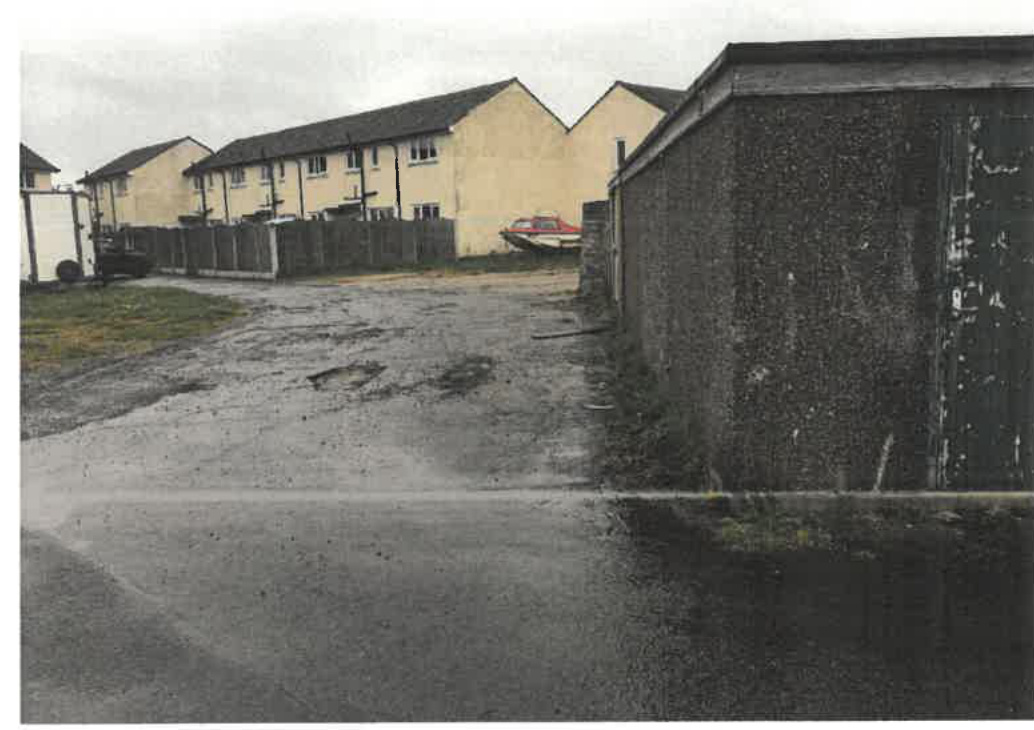 A photograph showing a paved yard area with a wooden fence and a large, dark block structure on the right, likely a disused garage, with residential buildings in the background.