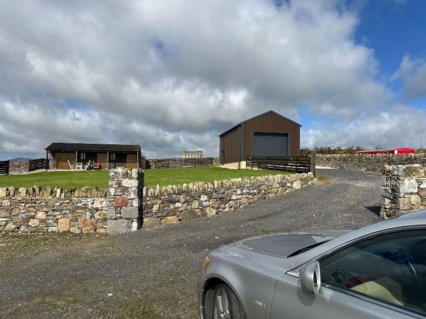 A photograph showing a rural property with dry stone walls and a gravel driveway. Two wooden outbuildings are visible, including a larger structure with a garage door.