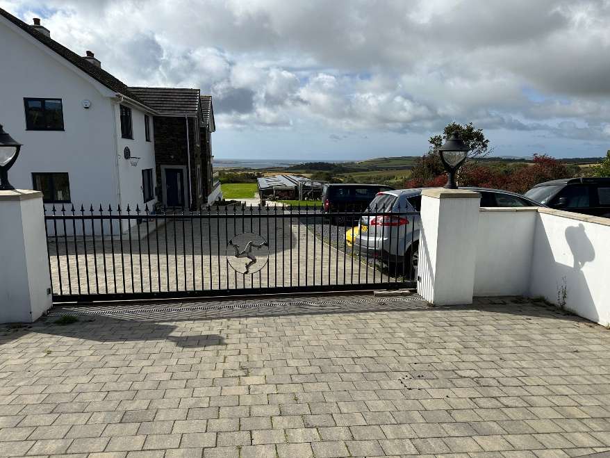 A photograph showing the front entrance of a white detached house with a paved driveway, black metal gate featuring a Manx triskelion, and a coastal view in the background.