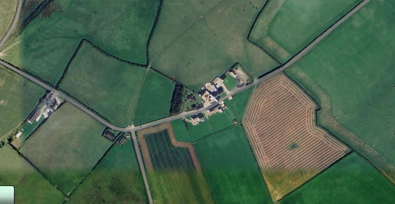 Aerial satellite view of a rural hamlet showing a cluster of buildings surrounded by green agricultural fields and roads.