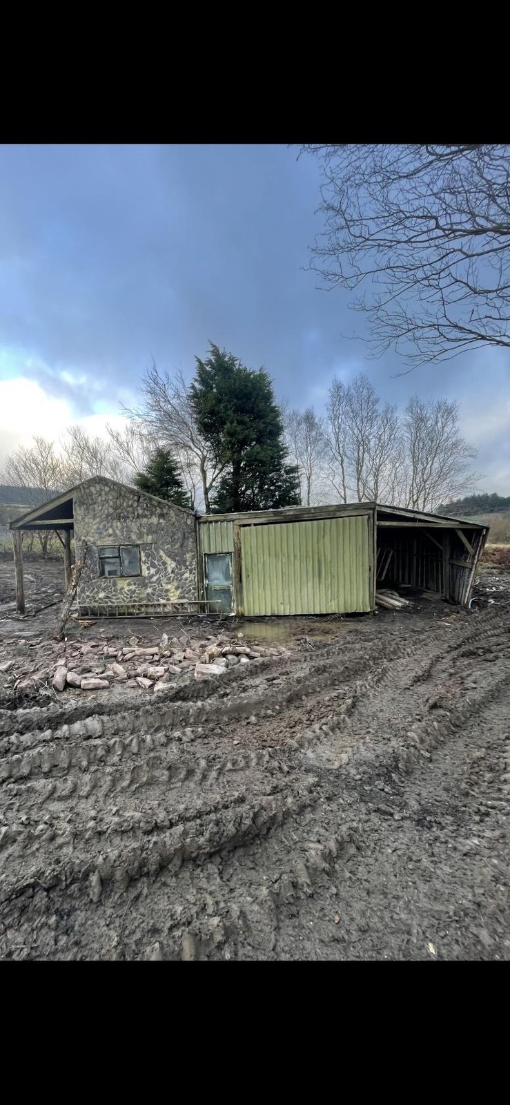 A photograph showing a dilapidated rural building consisting of a stone structure and a green corrugated metal extension, set against a muddy foreground with tire tracks.