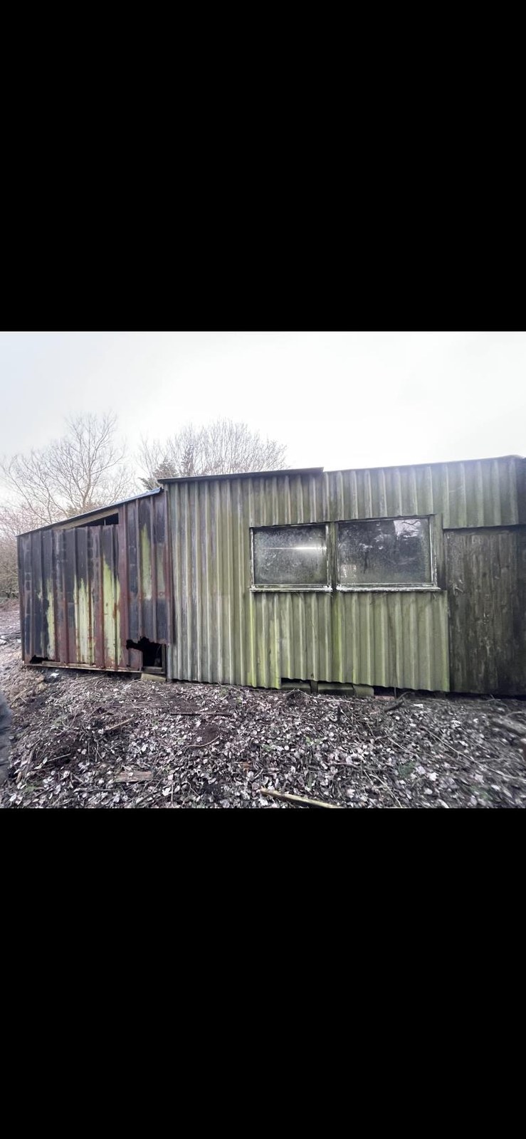 A photograph of a dilapidated, single-story corrugated metal outbuilding in a rural setting, showing significant weathering and partial structural damage.
