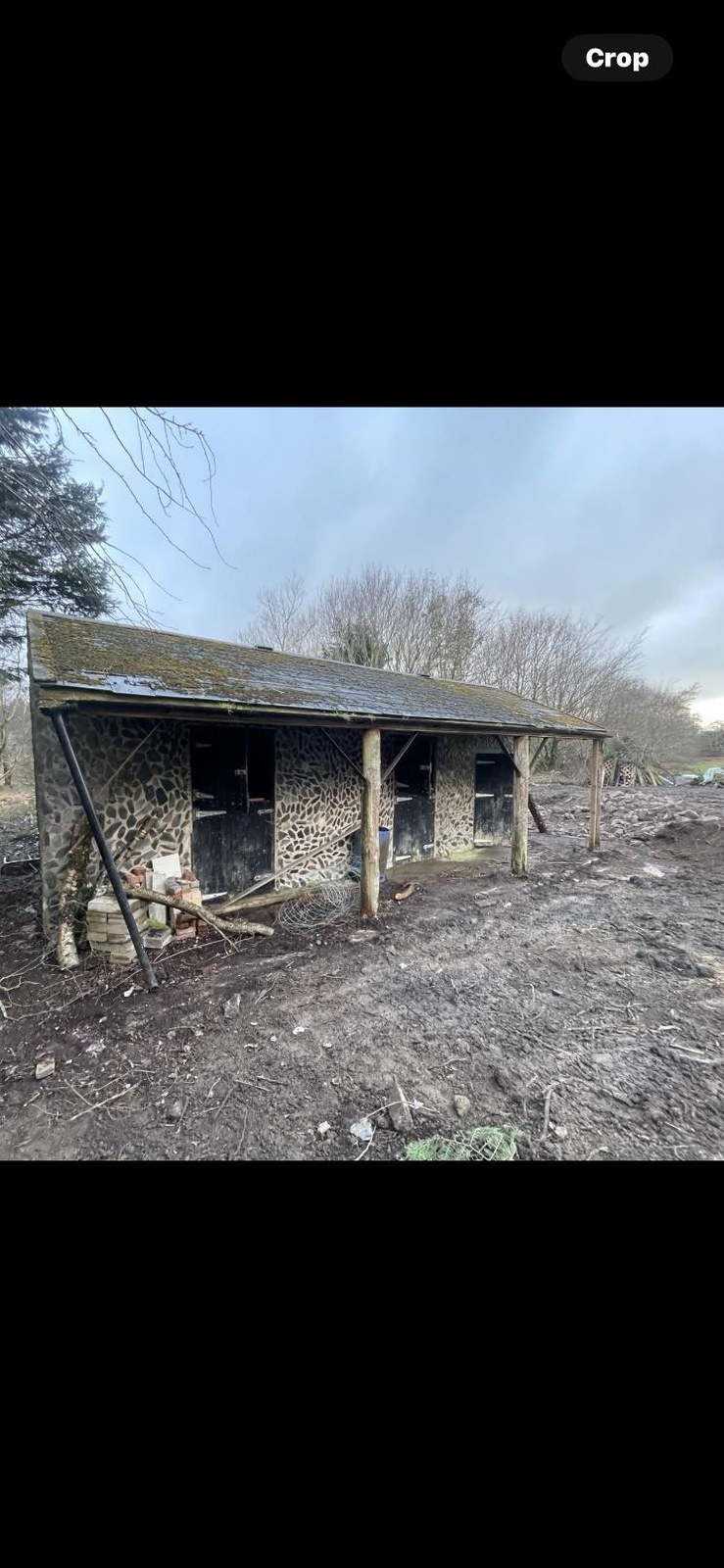 A photograph showing a dilapidated single-story stone building, likely a barn or agricultural outbuilding, in a rural setting with muddy ground.