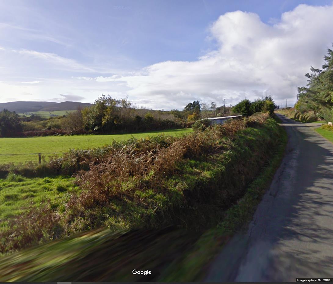 A Google Street View style photograph showing a rural road verge, green fields, and rolling hills in the background, likely depicting the site location.
