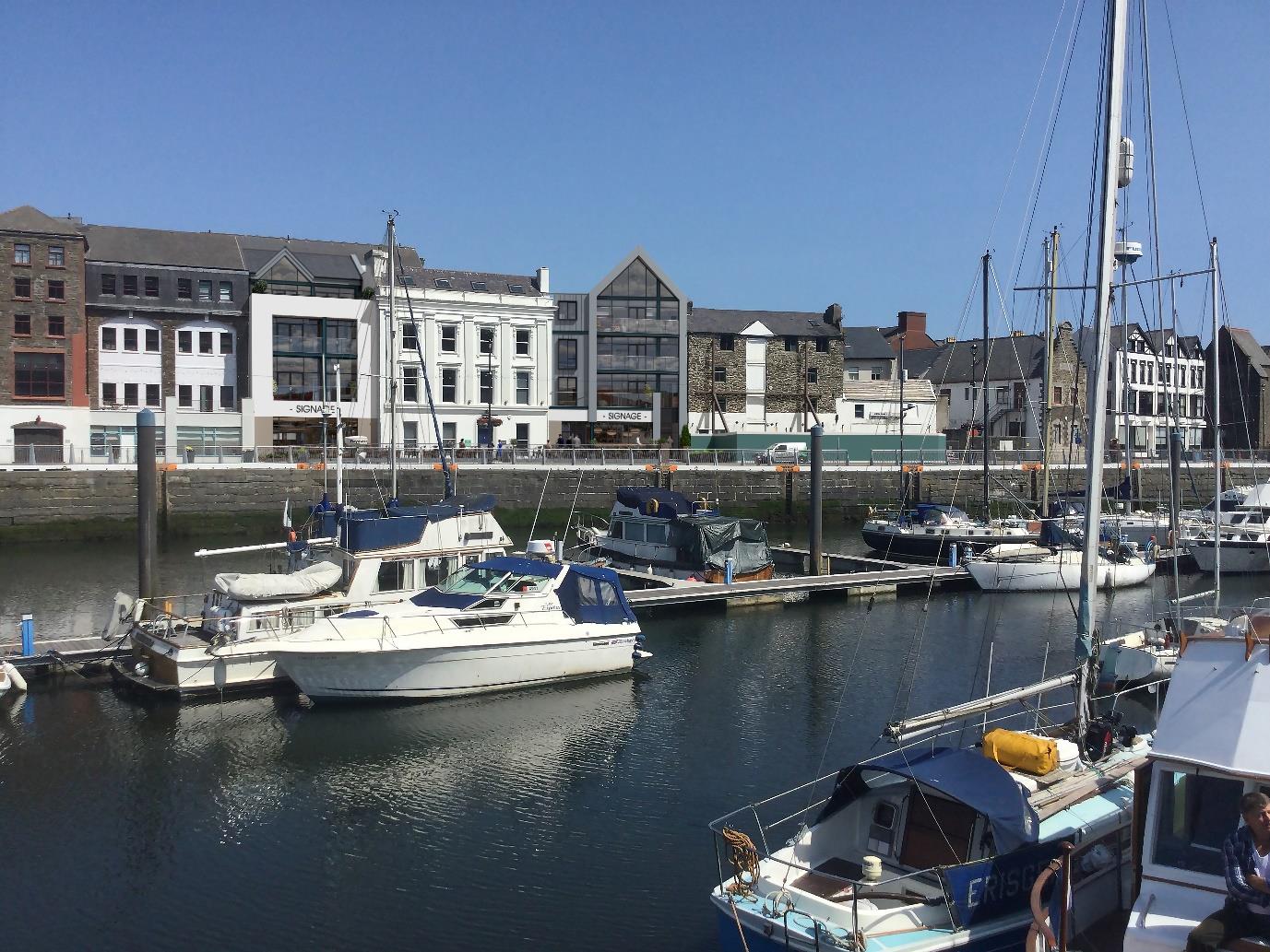 A photograph showing a harbor scene with several boats moored in the foreground and a row of mixed commercial and residential buildings along the waterfront in the background.