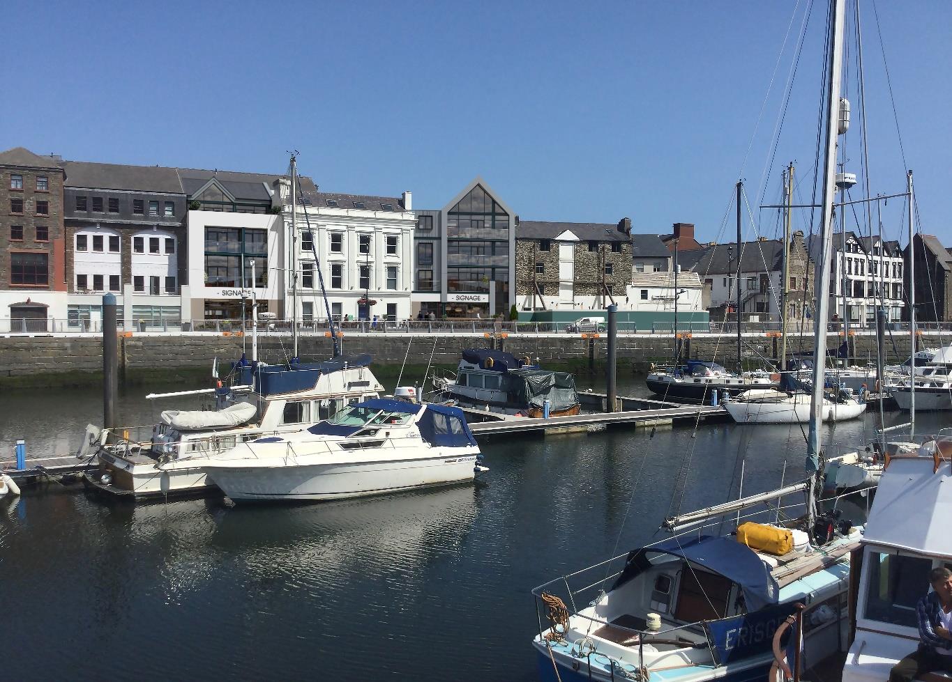 A sunny photograph showing a harbor with several boats moored in the foreground and a row of mixed commercial and residential buildings along the waterfront in the background.