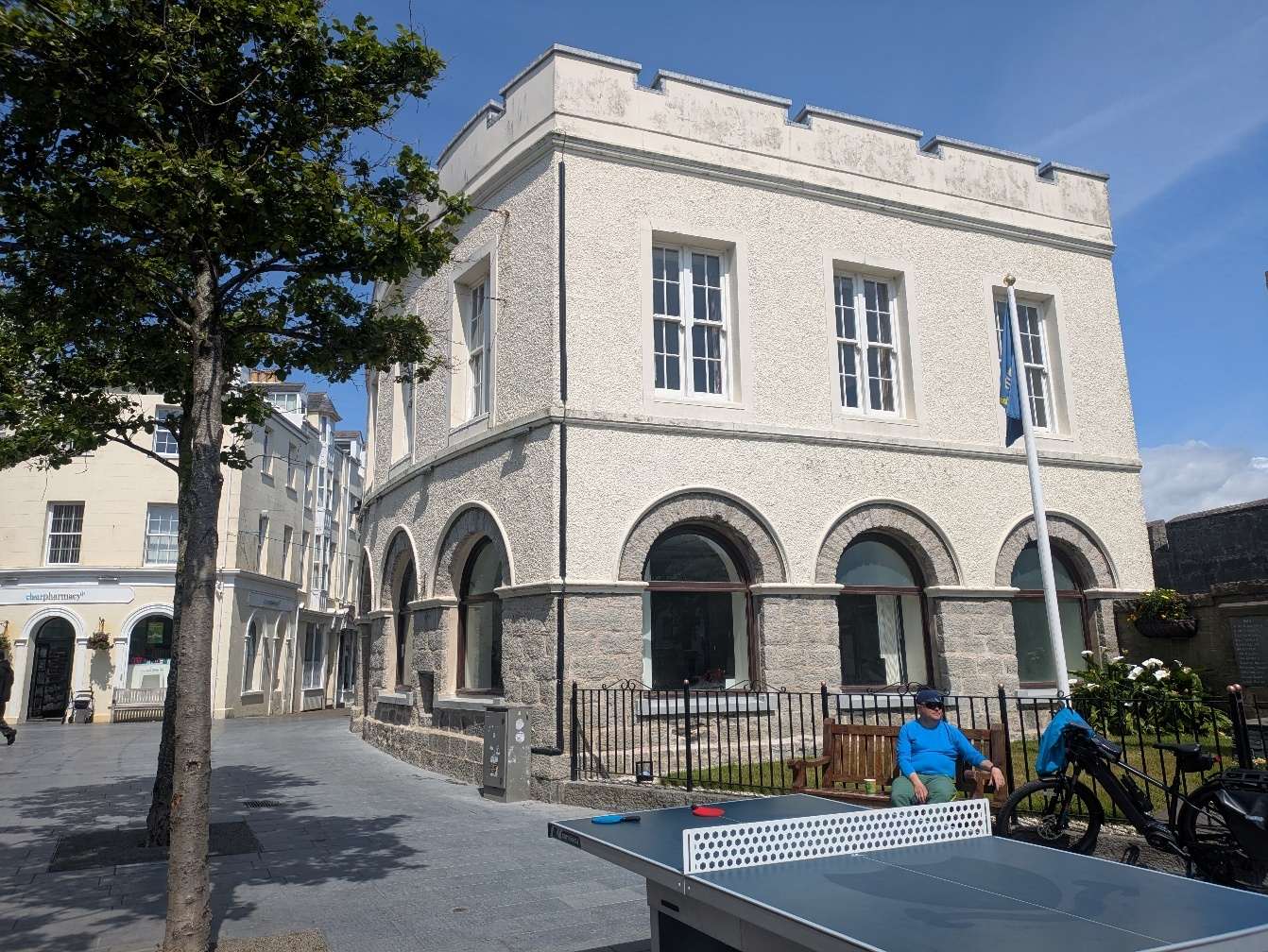 A photograph of a distinctive white two-story building with crenellated parapets and arched ground-floor windows, situated in a paved town square with a ping pong table in the foreground.