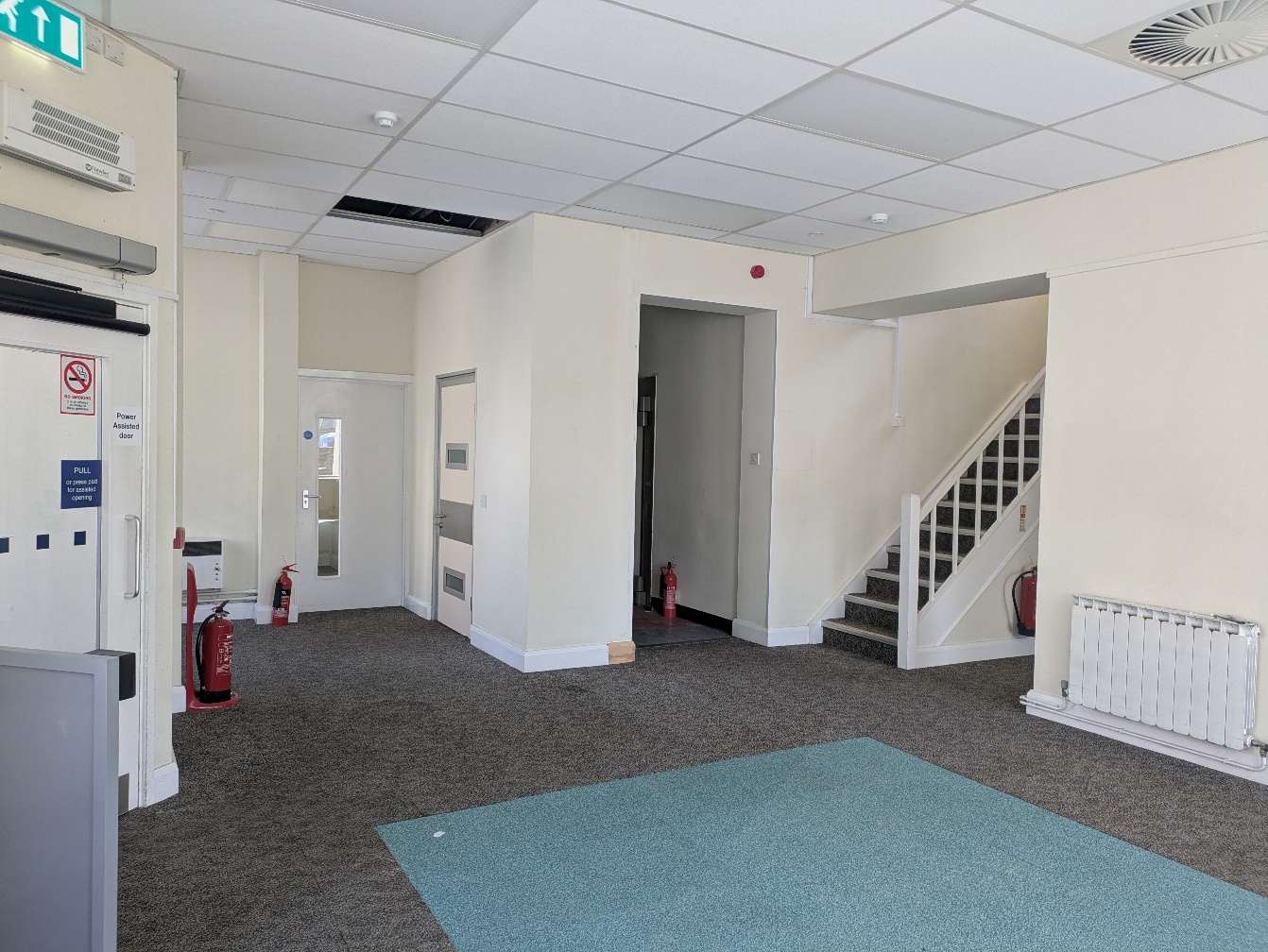 Interior photograph of a commercial building hallway featuring a staircase, power-assisted door, and fire safety equipment.