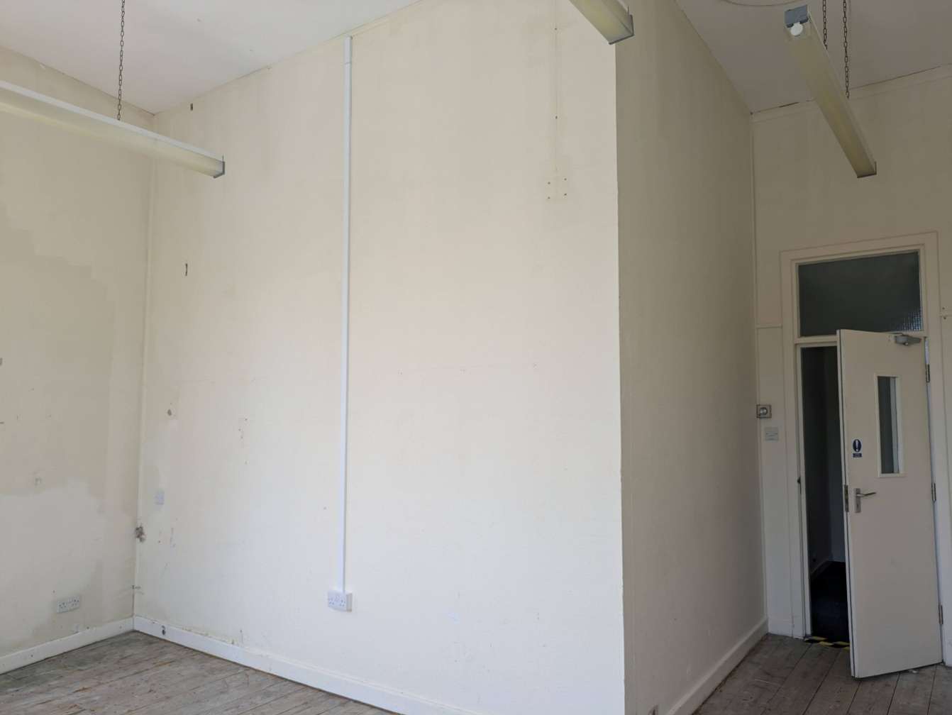 Interior photograph of an empty room featuring white walls, wooden flooring, and suspended fluorescent lights.