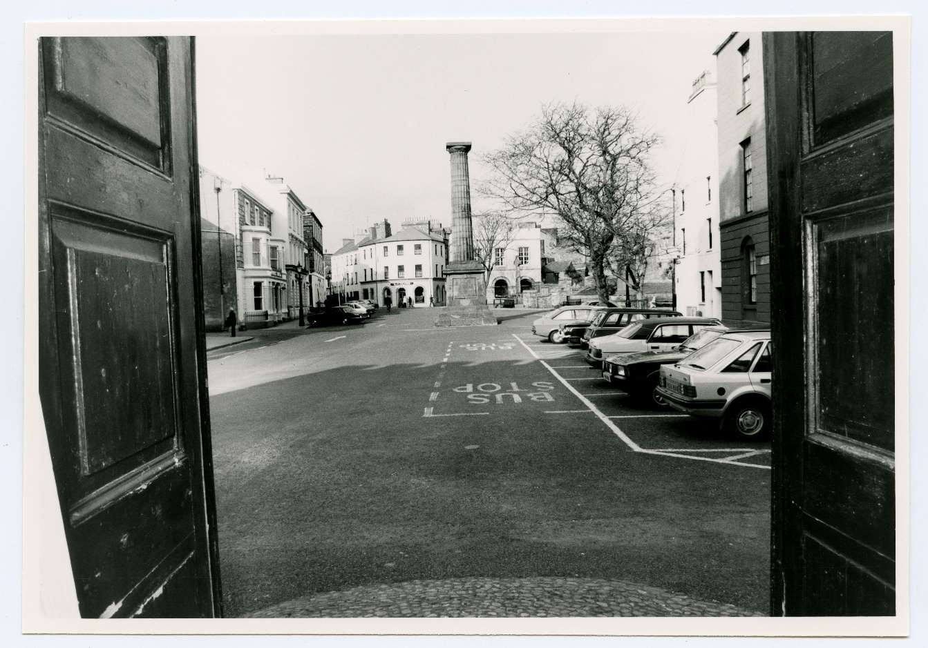 A black and white photograph taken from inside a building looking out through open doors onto a town street featuring a central column monument and parked cars.