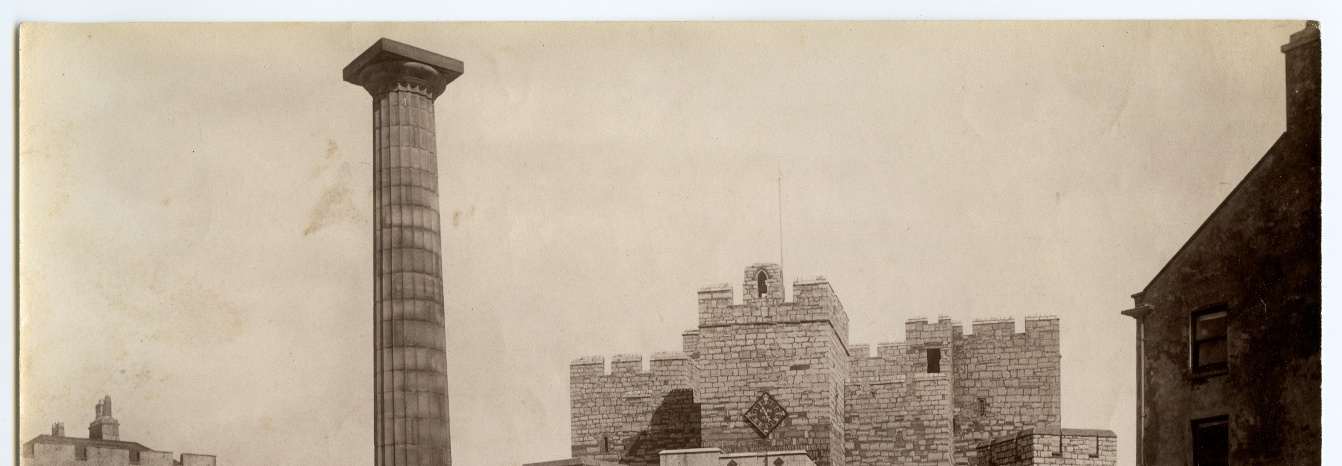 A sepia-toned historical photograph showing a tall fluted column monument standing next to a crenellated stone building.