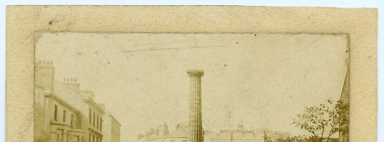 A vintage sepia-toned photograph depicting a street scene with terraced buildings on the left and a tall column monument in the center.