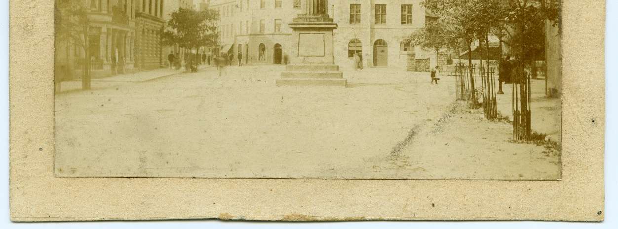 A vintage sepia-toned photograph depicting a town square with a central monument and surrounding historic buildings.