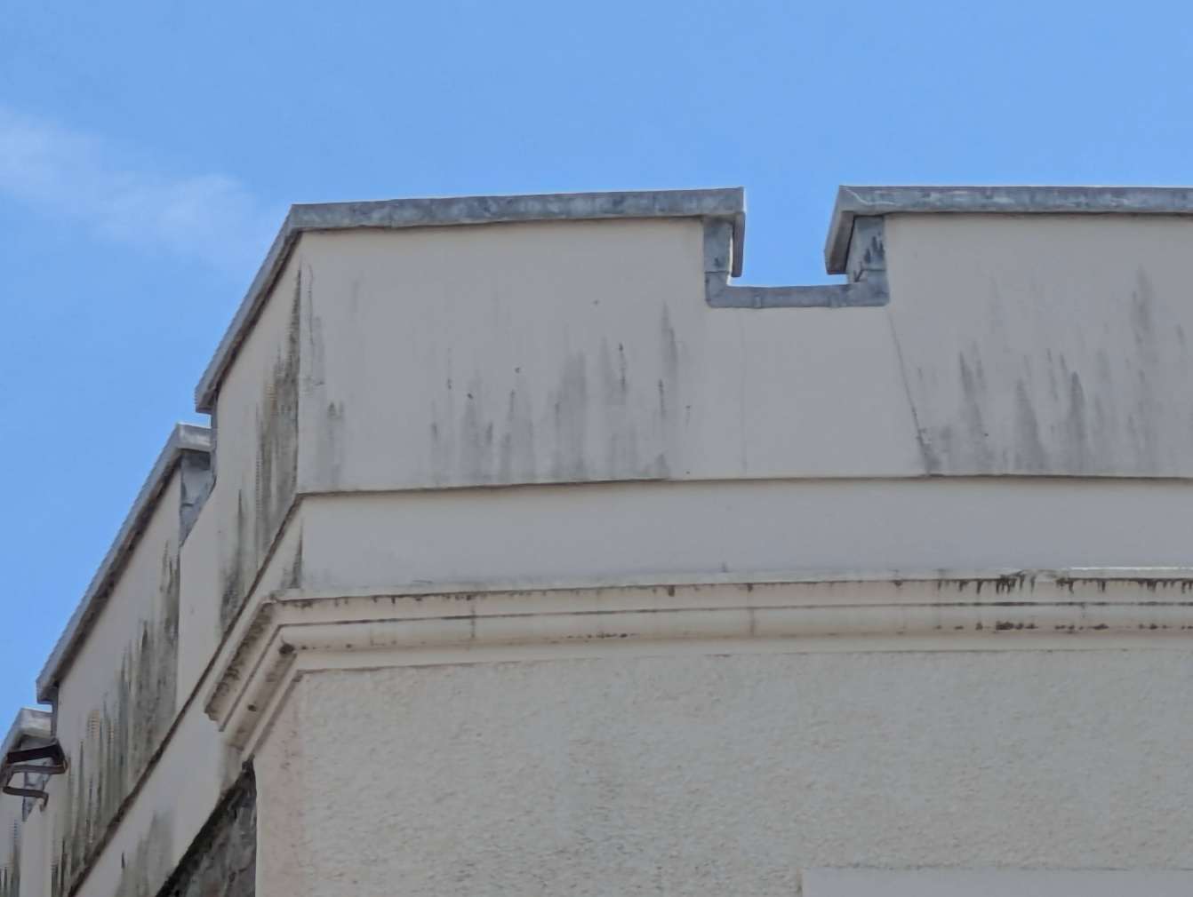 A close-up photograph showing the weathered upper facade and stone coping of a building parapet against a blue sky.
