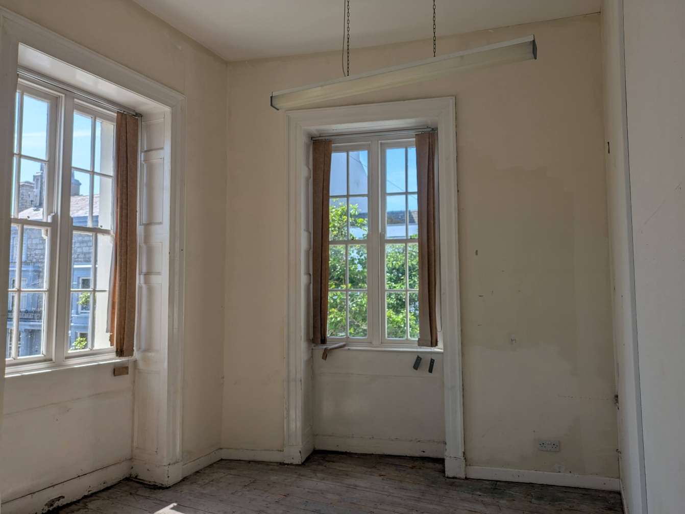 An interior photograph of an empty room featuring wooden floorboards and two white sash windows with curtains.